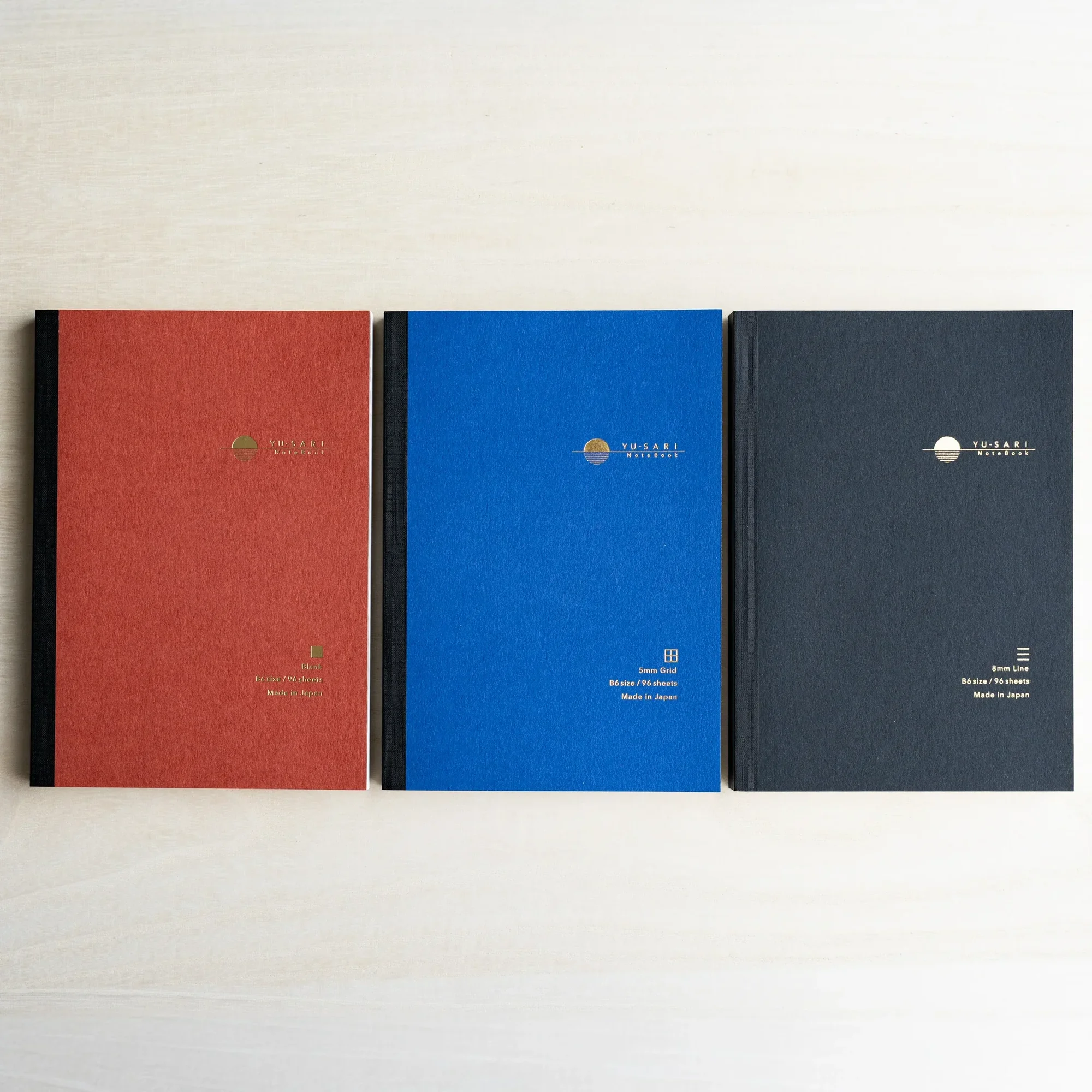 Three A6 YU-SARI notebooks lying side by side on wood, dark red Blank, black 8mm Line, and blue 5mm Grid with gold logos