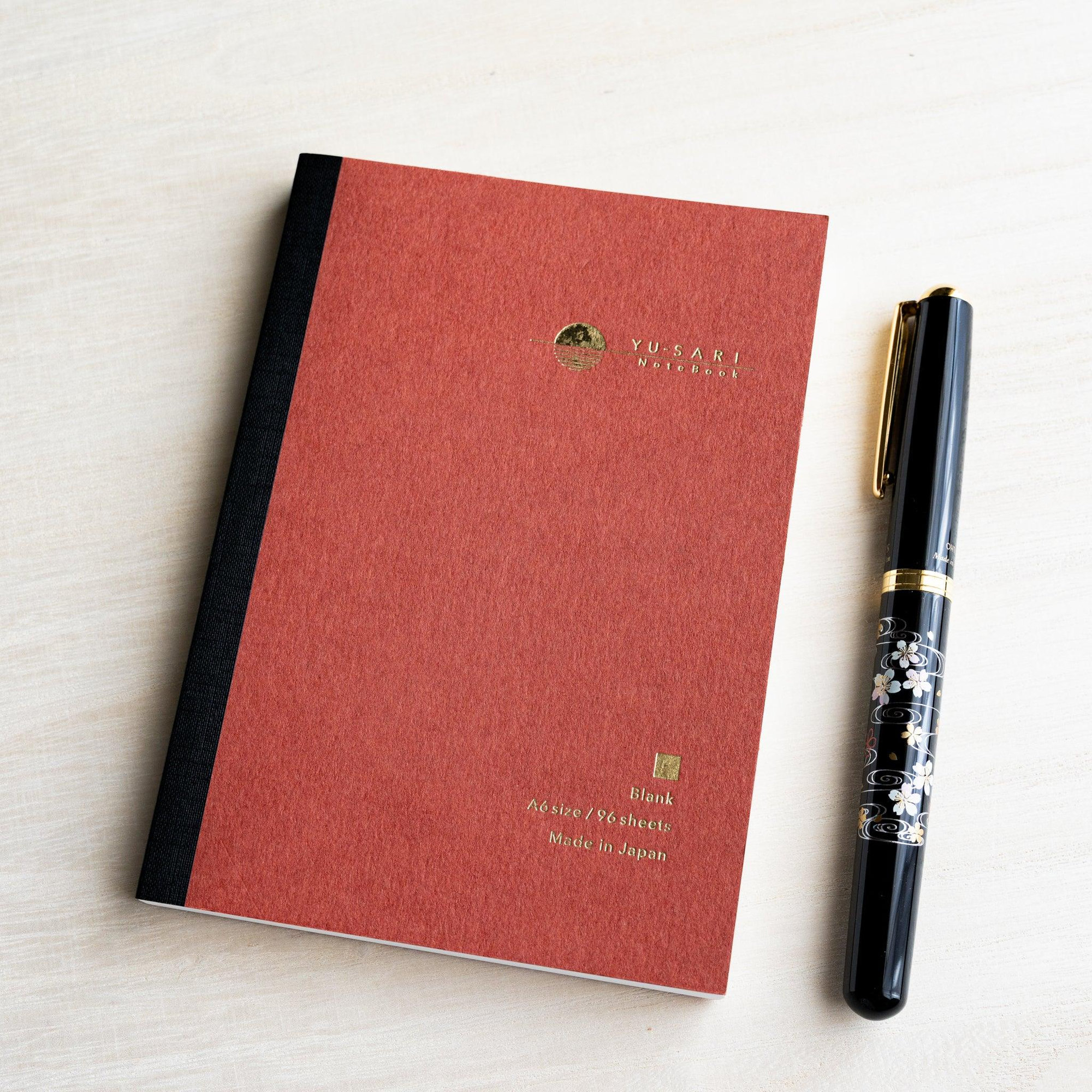 Dark red A6 YU-SARI blank notebook on wood beside a black lacquer maki-e fountain pen, gold Blank, A6 size, 96 sheets text