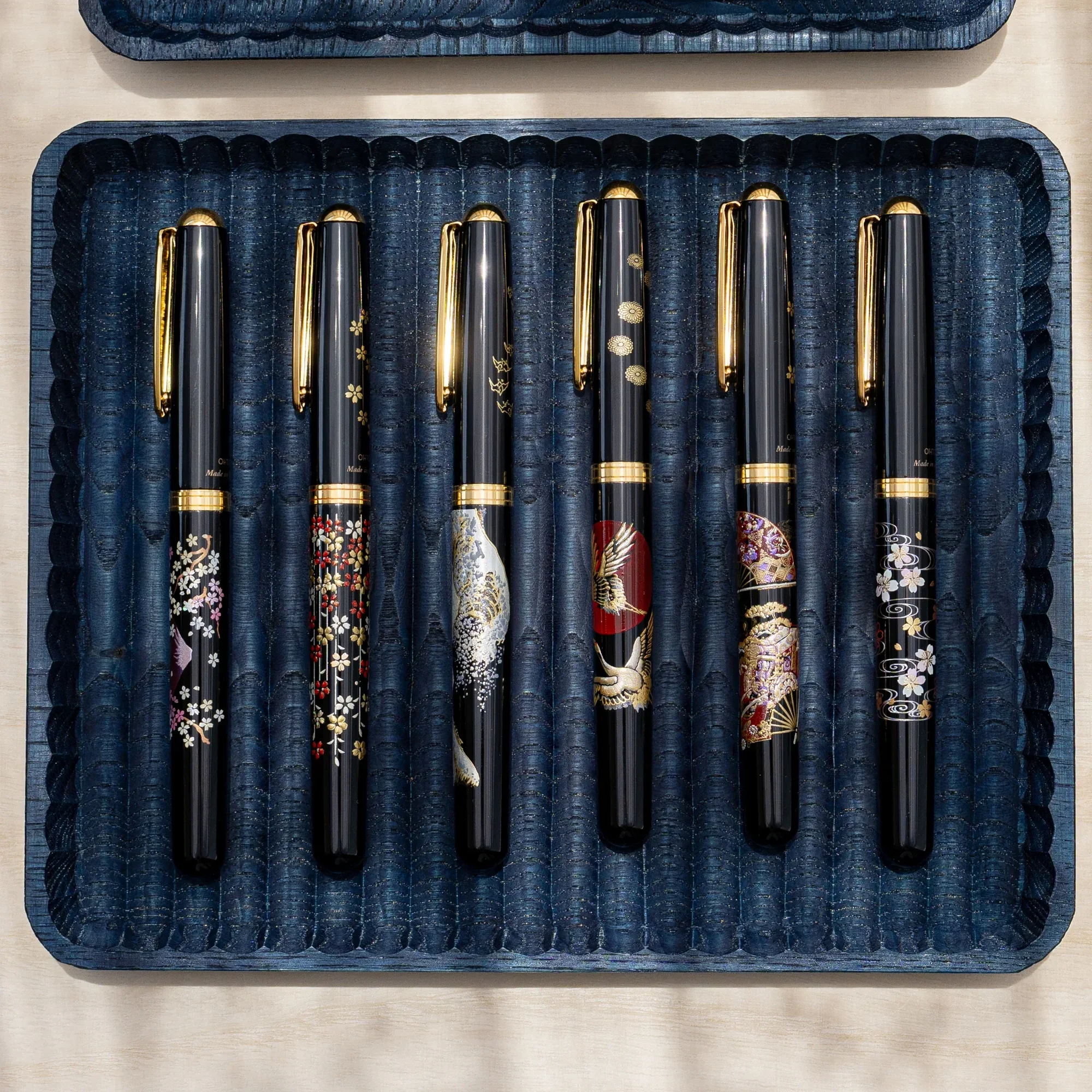 Indigo dyed wide chestnut pen tray from above holding six black fountain pens with gold and colored designs